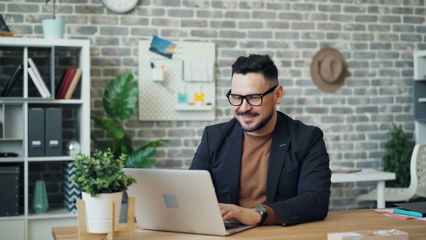 Man with beard smiles while working on a laptop at a desk.